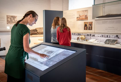 A woman is using the digital display, while in the background, another woman and a young girl are reading the exhibits in the exhibition at Sønderborg Castle.