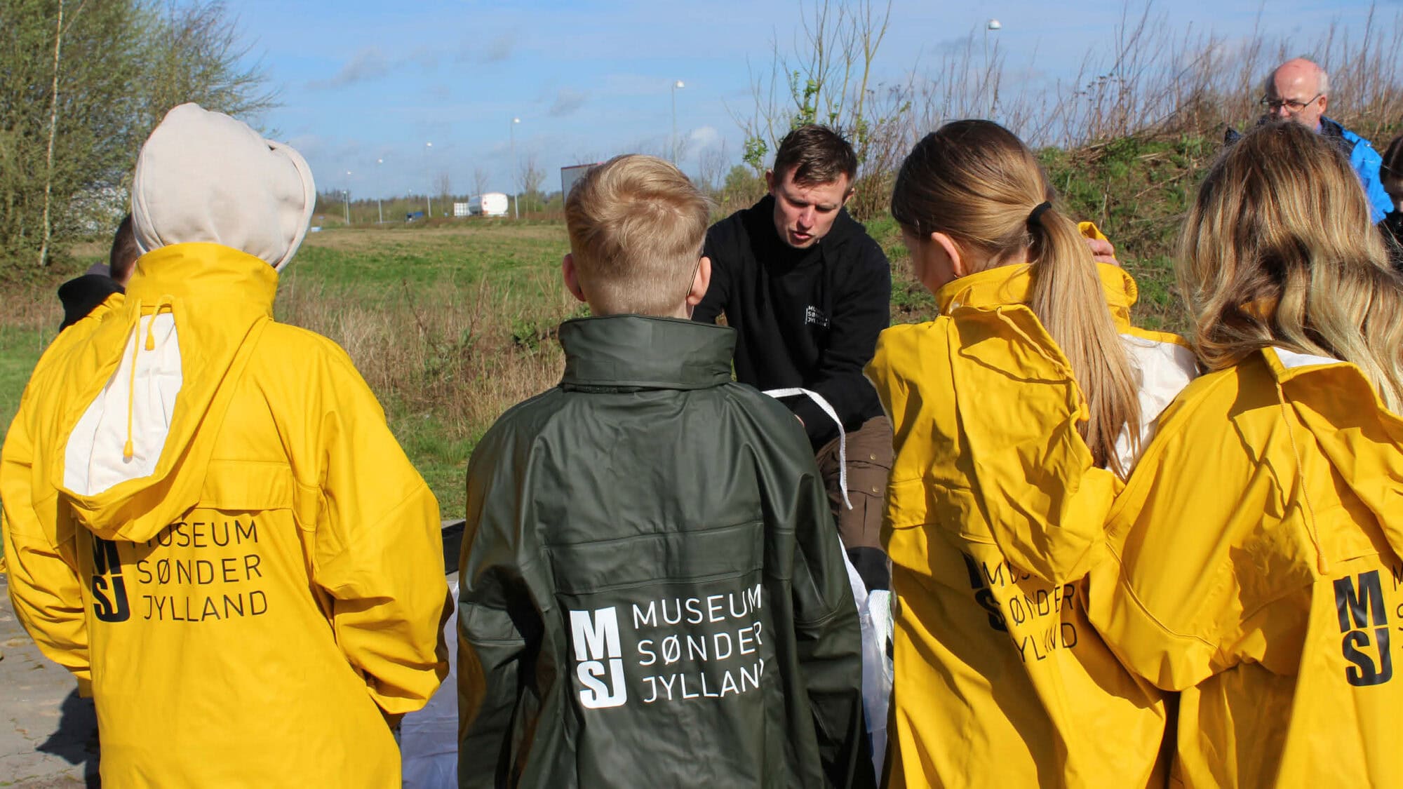 Børn står i regnfrakker klar til at slemme jord og finde genstande fra en arkæologisk udgravning.