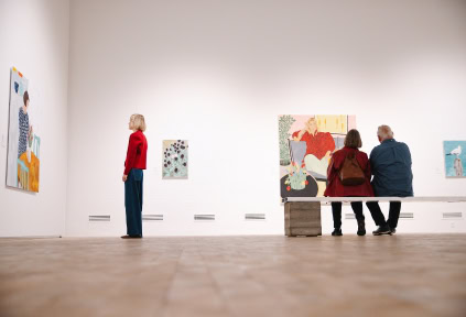 A couple is sitting on a bench, and a young woman stands next to them. They are all admiring artwork by Anette Abrahamsson at the Art Museum in Tønder