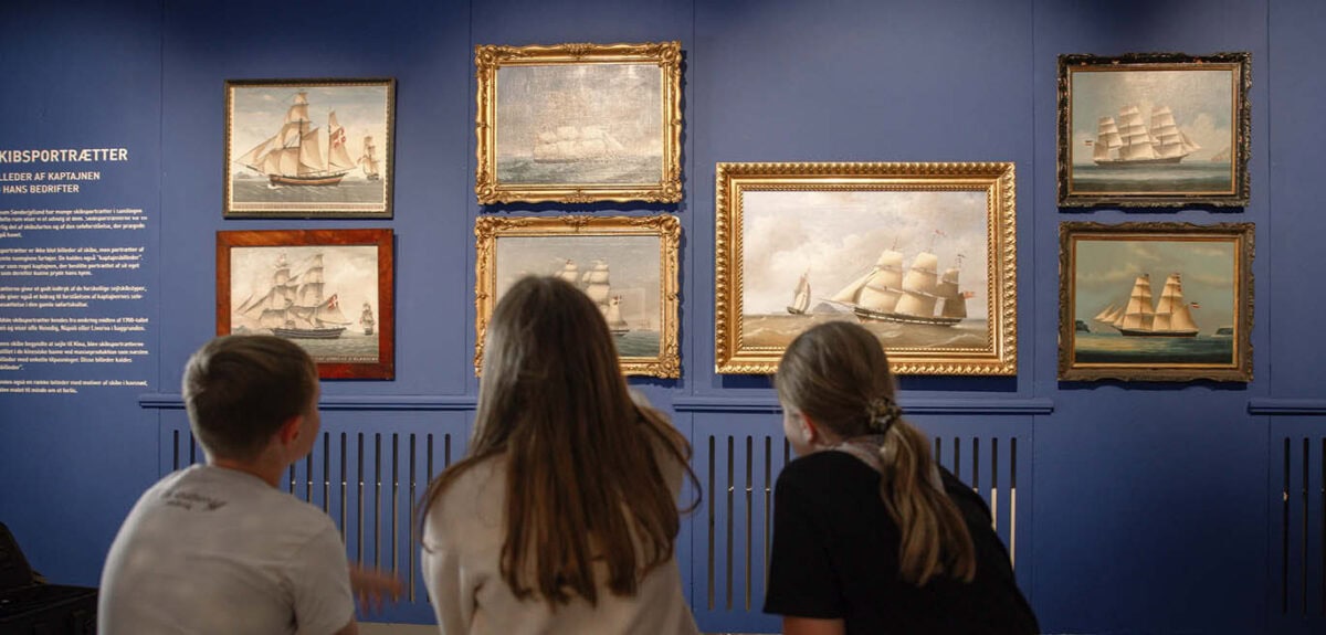 To piger og en dreng sidder i et blåt rum fyldt med skibsportrætter