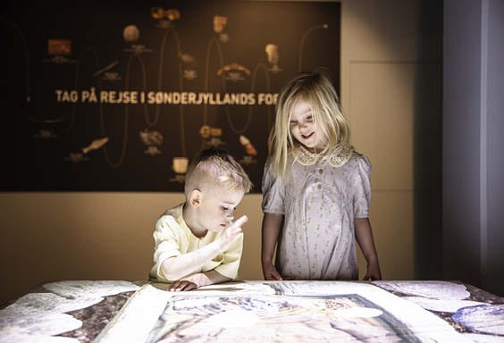 En dreng og pige står ved en et stort bord, hvor drengen skal til at trykke på noget på Arkæologi Haderslev.