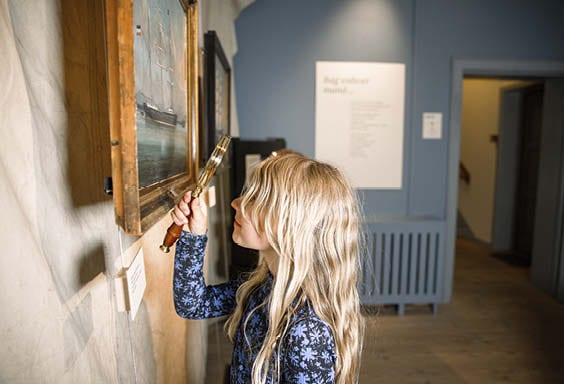 Ung pige står med en lup vendt og undersøger et maleri på Kulturhistorie Aabenraa